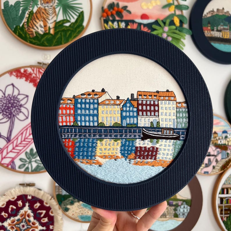 Puede incluir: Un aro de bordado azul marino con un fondo blanco que presenta una ciudad bordada en colores con edificios y un r&iacute;o. La ciudad est&aacute; rodeada por un borde blanco.