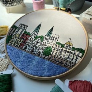 May include: Embroidered hoop art depicting a cityscape with a church, buildings, and a river. The buildings are in shades of brown, red, and white, with green trees and a blue river. The hoop is made of wood and has a white background.