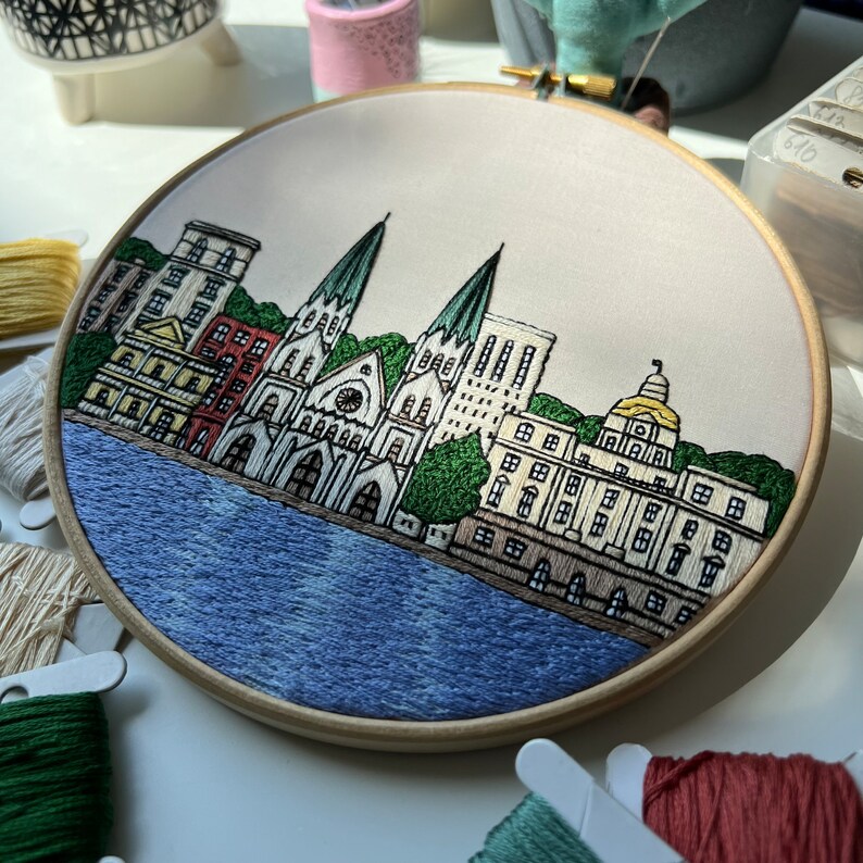 May include: Embroidered hoop art depicting a cityscape with a church, buildings, and a river. The buildings are in shades of brown, red, and white, with green trees and a blue river. The hoop is made of wood and has a white background.