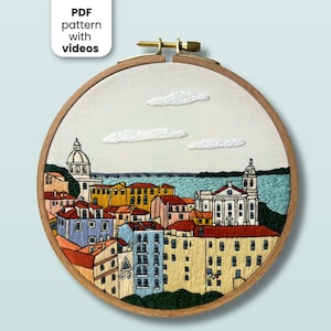 May include: A circular wooden embroidery hoop containing a hand-stitched scene of a coastal city with colourful buildings, a church with a dome, a bell tower, and a body of water under a pale sky with three white clouds. Text reads PDF pattern with videos.