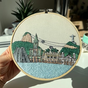 Puede incluir: Arte de bordado en aro que representa una ciudad con una iglesia, edificios, una monta&ntilde;a y una estatua en una colina. La escena est&aacute; representada en tonos de verde, marr&oacute;n y azul.