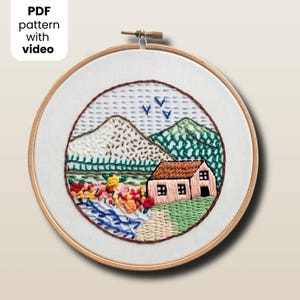 May include: Embroidered landscape art in a wooden hoop. The scene includes a house, mountains, flowers, and a river, all stitched with colorful thread. The text "PDF pattern with video" is in the upper left corner.