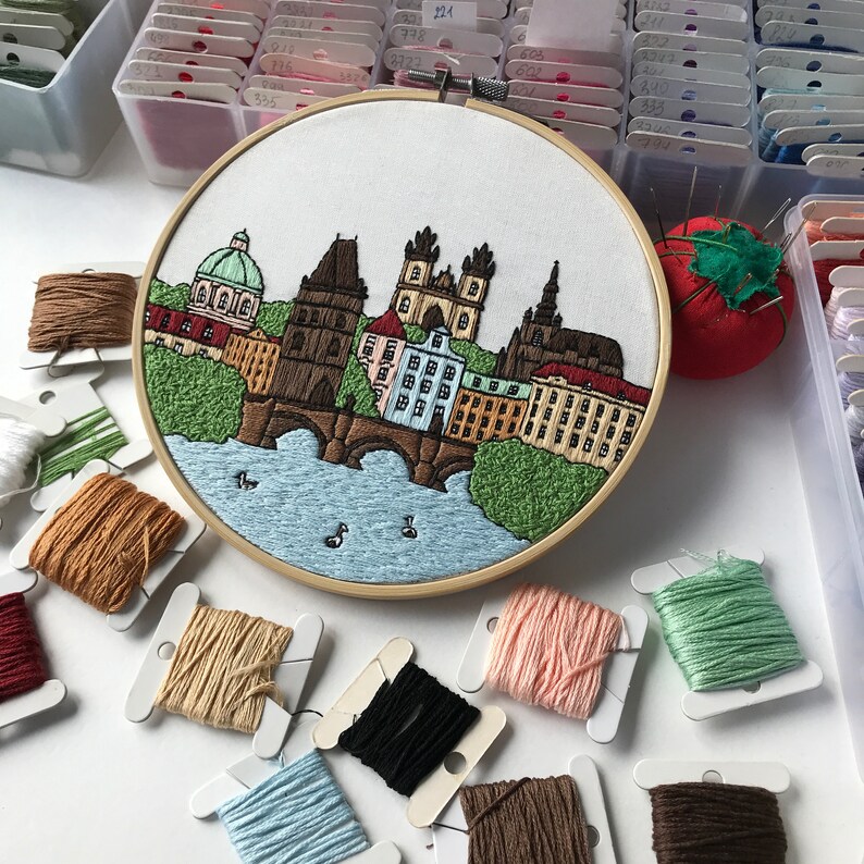 May include: A close-up of a completed embroidery hoop with a colorful depiction of the Charles Bridge in Prague, Czech Republic. The hoop is surrounded by various colors of embroidery floss in small plastic spools.