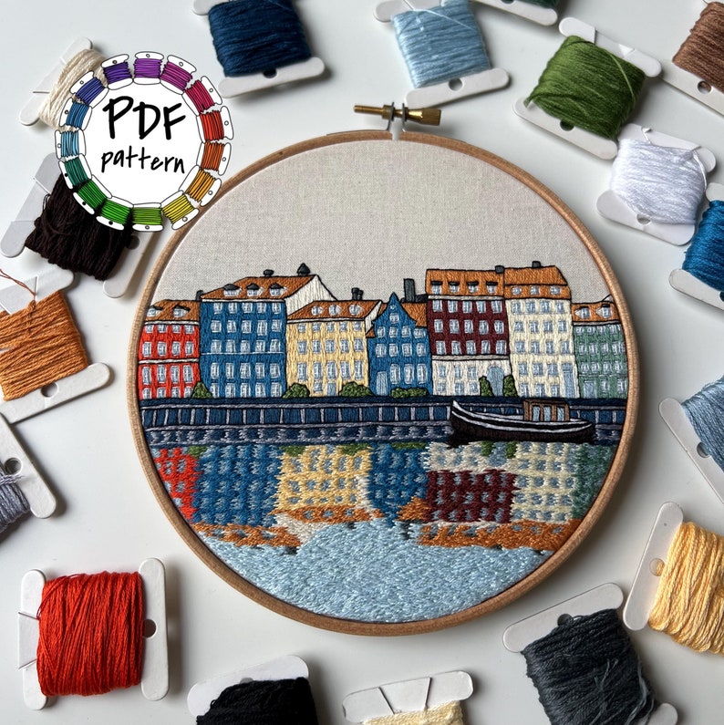 Puede incluir: Arte de bordado en aro que representa una ciudad con edificios coloridos a lo largo de una costa. Los edificios son en tonos de rojo, amarillo, azul y verde. El agua es de color azul claro con un reflejo de los edificios. El aro es de madera y tiene un fondo blanco.