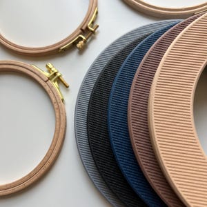 May include: Six embroidery hoops in various colors, including gray, black, blue, brown, and beige. The hoops are stacked on top of each other and have a textured surface.