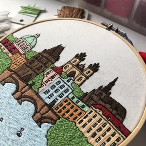 May include: Embroidered cityscape with a river and bridge, featuring buildings in shades of brown, red, and beige, with green trees and a blue sky. The embroidery is done on a white fabric and is framed in a wooden hoop.