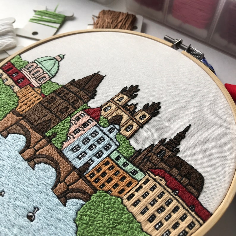 May include: Embroidered cityscape with a river and bridge, featuring buildings in shades of brown, red, and beige, with green trees and a blue sky. The embroidery is done on a white fabric and is framed in a wooden hoop.