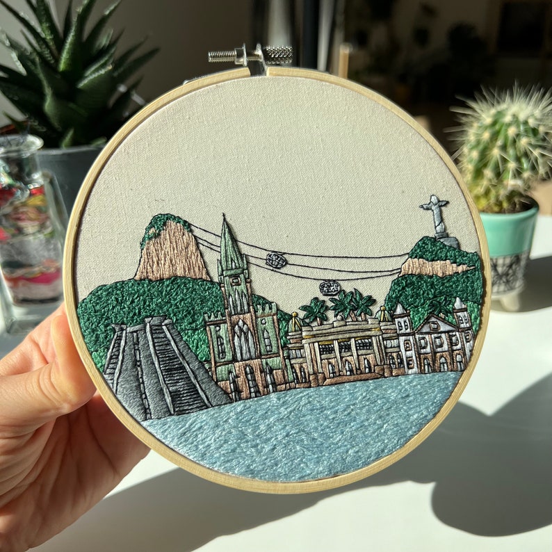Puede incluir: Bordado que representa una ciudad con una iglesia, edificios y una monta&ntilde;a en el fondo. La obra est&aacute; enmarcada en un aro de madera y presenta una gama de colores azules y verdes.