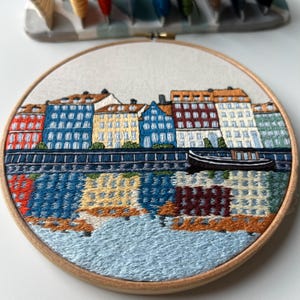 Puede incluir: Arte de bordado en aro que representa una ciudad con edificios coloridos a lo largo de una costa. Los edificios son en tonos de azul, amarillo, rojo y verde. Un peque&ntilde;o barco est&aacute; en el agua frente a los edificios.