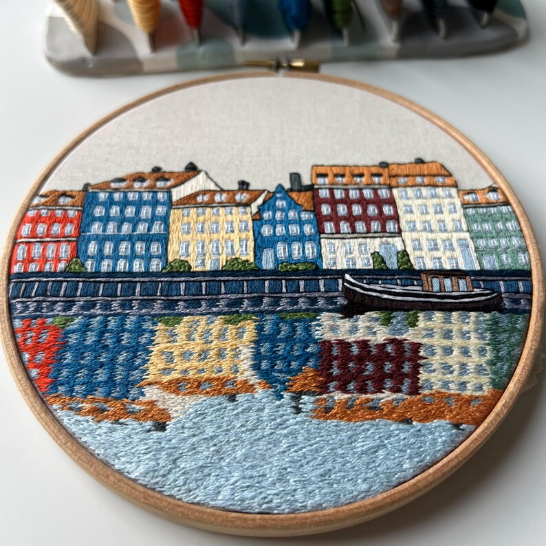 Puede incluir: Arte de bordado en aro que representa una ciudad con edificios coloridos a lo largo de una costa. Los edificios son en tonos de azul, amarillo, rojo y verde. Un peque&ntilde;o barco est&aacute; en el agua frente a los edificios.
