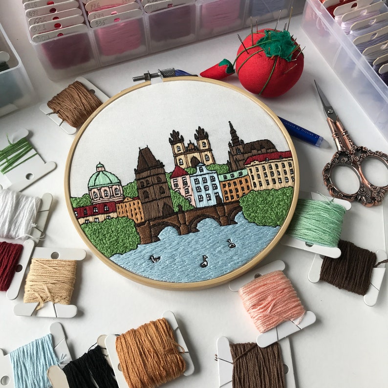 May include: A close-up of a completed embroidery hoop with a colorful depiction of a cityscape, including buildings, a bridge, and trees. The hoop is surrounded by various colors of embroidery floss.