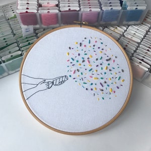 May include: Embroidered hoop art featuring a hand holding a party popper with colorful confetti exploding from it. The confetti is made of different colored embroidery floss.