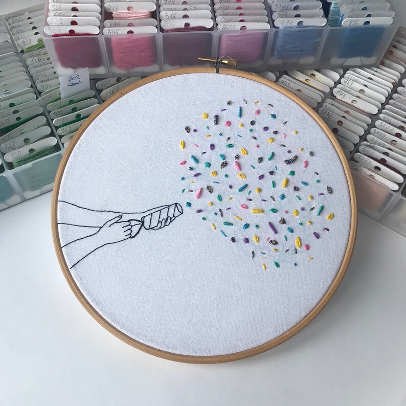 May include: Embroidered hoop art featuring a hand holding a party popper with colorful confetti exploding from it. The confetti is made of different colored embroidery floss.