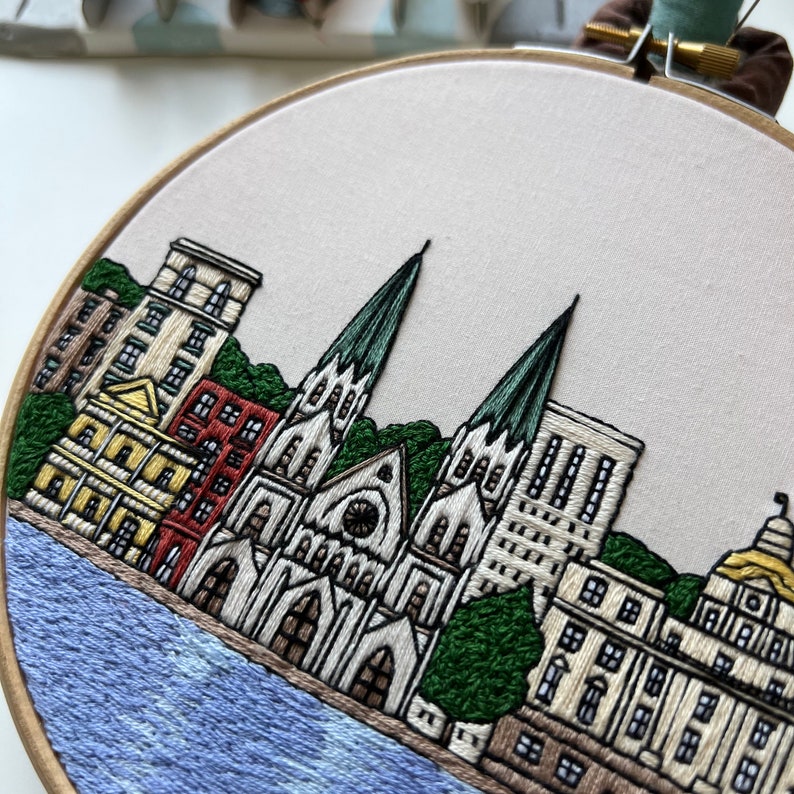 May include: Embroidered cityscape with a large church building in the center, surrounded by smaller buildings and trees. The scene is rendered in shades of brown, green, blue, and white on a beige fabric.