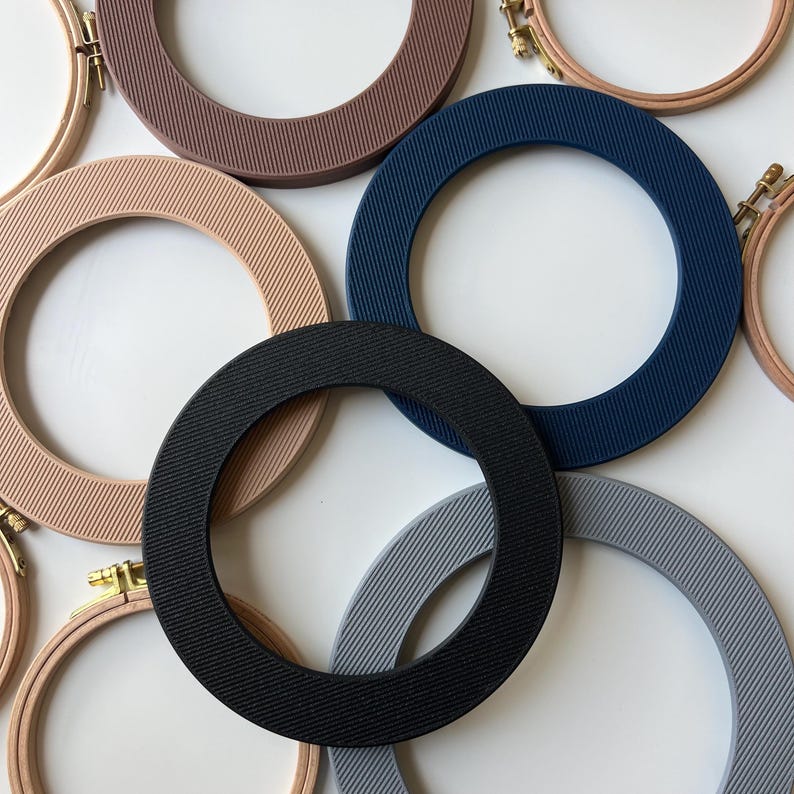 May include: Assortment of embroidery hoops in various colors, including beige, dark blue, gray, and black. The hoops are made of plastic and have a ribbed texture.
