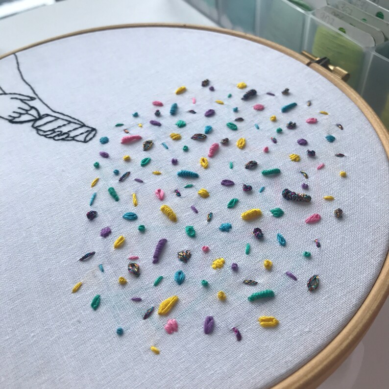 May include: A white embroidery hoop with a hand-stitched design of a hand holding a container pouring out colorful confetti. The confetti is made of various colors of embroidery floss, including yellow, pink, blue, green, and purple.