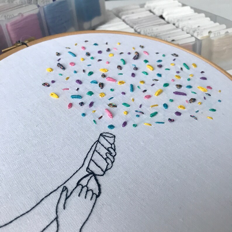 May include: Embroidered hoop art featuring a hand holding a party popper with colorful confetti exploding from it. The confetti is made of various colors of embroidery floss, including pink, yellow, green, purple, and blue.