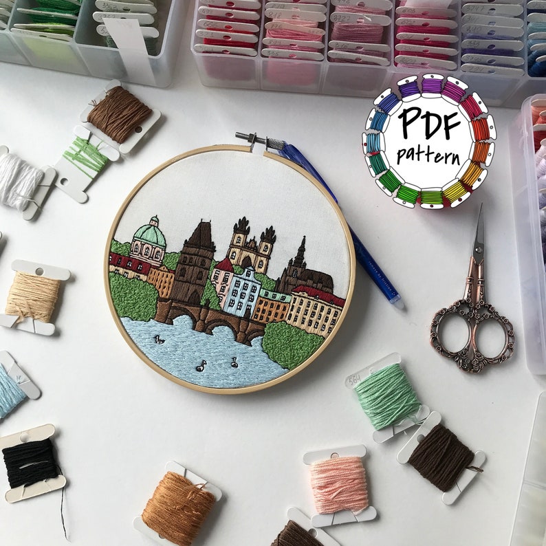 May include: Embroidered hoop art featuring a cityscape of Prague, Czech Republic. The scene includes the Charles Bridge, buildings, and a river. The hoop is surrounded by embroidery floss spools in various colors.