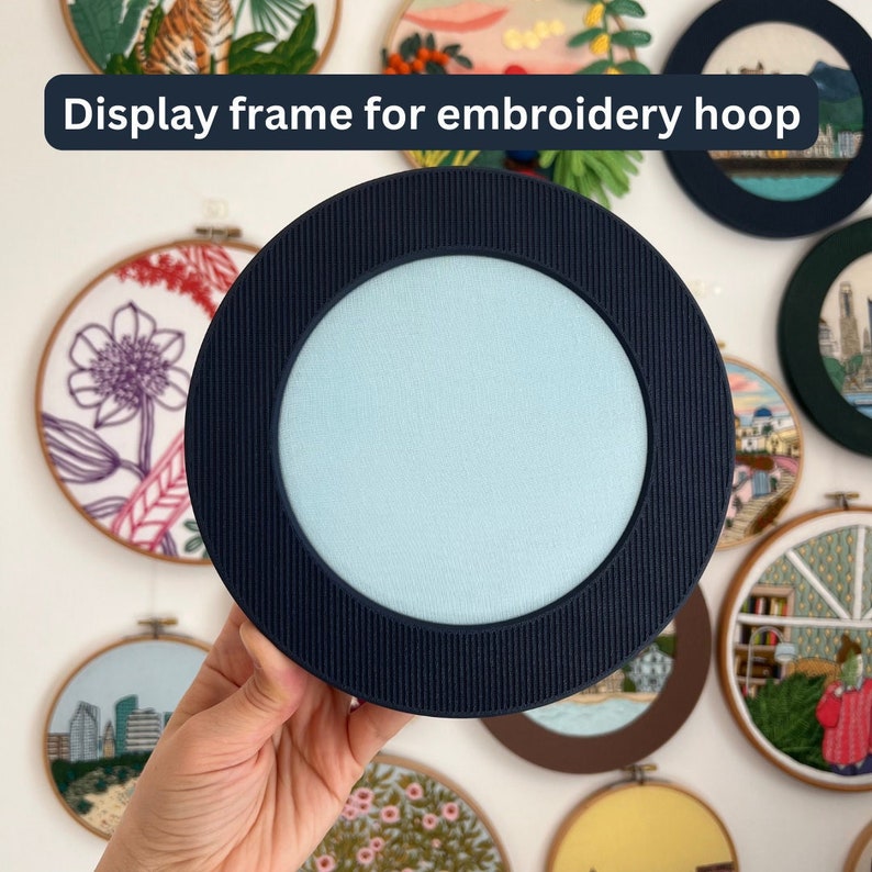 A circular 3D-printed display frame showcases an embroidery hoop with blue fabric inside, held in hand againt the wall. The minimalist frame surrounds the hoop evenly, offering a clean, modern look ideal for wall decor. It serves as a decorative and functional solution for hanging embroidery art without visible nails or hardware.