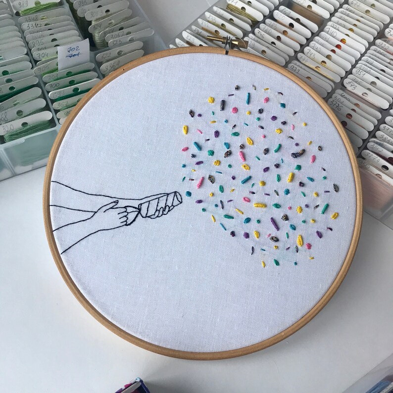 May include: A white embroidery hoop with a hand-stitched design of a hand holding a party popper with confetti exploding out of it. The confetti is in various colors, including pink, blue, yellow, green, and purple.