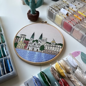 May include: Embroidered hoop art depicting a cityscape with a waterfront and several buildings, including a church with two towers. The buildings are rendered in shades of beige, red, and green, with a blue water scene.