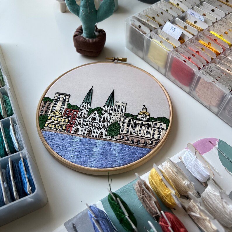 May include: Embroidered hoop art depicting a cityscape with a waterfront and several buildings, including a church with two towers. The buildings are rendered in shades of beige, red, and green, with a blue water scene.