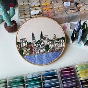May include: Embroidered cityscape with a blue water scene. The embroidery features a row of buildings with two tall churches in the center. The buildings are in shades of red, yellow, and brown. The embroidery is framed in a wooden hoop.
