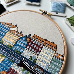 Puede incluir: Arte de bordado en aro que representa una ciudad con edificios coloridos y un peque&ntilde;o barco en el agua. Los edificios son en tonos de amarillo, naranja, rojo, verde y azul. El agua es de un azul claro con un patr&oacute;n de olas blancas y azules.