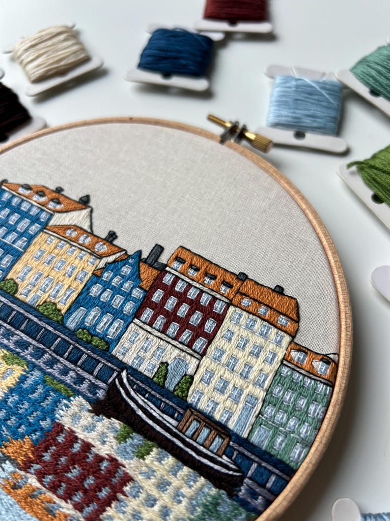 Puede incluir: Arte de bordado en aro que representa una ciudad con edificios coloridos y un peque&ntilde;o barco en el agua. Los edificios son en tonos de amarillo, naranja, rojo, verde y azul. El agua es de un azul claro con un patr&oacute;n de olas blancas y azules.