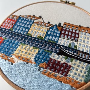 Puede incluir: Arte de bordado en aro que representa una ciudad con edificios coloridos a lo largo de una costa. Los edificios son en tonos de rojo, azul, amarillo y marr&oacute;n. El agua se representa en tonos de azul y verde, con un peque&ntilde;o barco en primer plano.