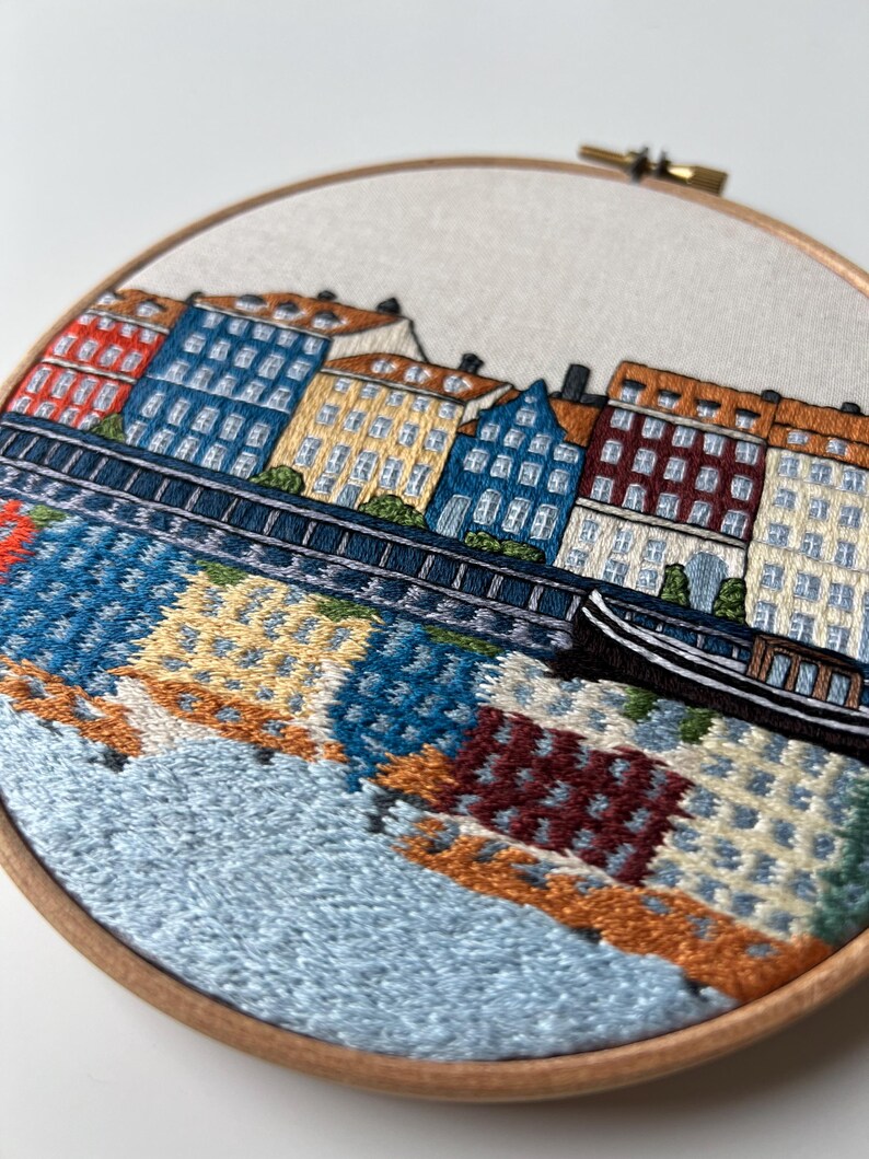 Puede incluir: Arte de bordado en aro que representa una ciudad con edificios coloridos a lo largo de una costa. Los edificios son en tonos de rojo, azul, amarillo y marr&oacute;n. El agua se representa en tonos de azul y verde, con un peque&ntilde;o barco en primer plano.