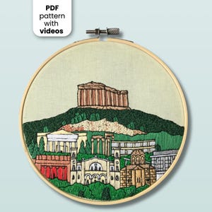 May include: An embroidery hoop containing a fabric panel with a hand-stitched depiction of the Acropolis and surrounding Athenian architecture. The design features tan, green, and red thread work. A label reads PDF pattern with videos.