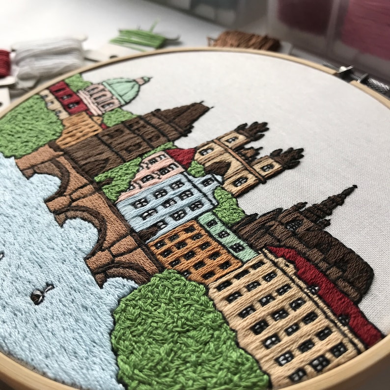 May include: Embroidered cityscape with a brown bridge, buildings with windows, and green trees. The embroidery is on a white fabric stretched over a wooden hoop.