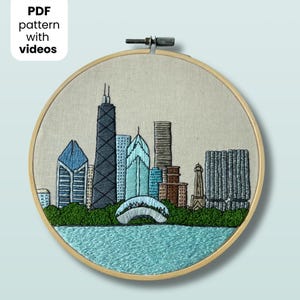 May include: A circular wooden embroidery hoop containing a fabric panel with a hand-stitched depiction of the Chicago skyline. The design features various skyscrapers in shades of blue, gray, and brown, set against a green park and blue water.