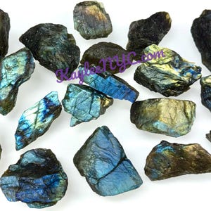 May include: A collection of raw labradorite stones in various shapes and sizes. The stones display a mix of dark gray, blue, and gold colors, with iridescent flashes. The rough texture of the stones is visible, and they are arranged on a white surface.