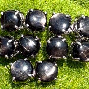 Könnte beinhalten: Eine Sammlung schwarzer Sternsaphir-Ringe mit silberfarbenen Fassungen. Die runden, polierten Edelsteine zeigen einen Sterneffekt. Die Ringe sind auf einem Bett aus grünem Gras angeordnet, was einen Kontrast von Farben und Texturen erzeugt.