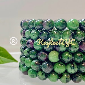 May include: Stack of green and black gemstone bracelets with a pink hue. The bracelets are made of round beads and are stacked on top of each other. The text "Kaylee NYC" is visible on the bracelets.