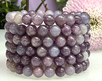 Puede incluir: Una pila de seis pulseras de cuentas hechas de piedras pulidas, redondas y lisas, en varios tonos de morado, lavanda y gris claro. Cada pulsera consta de cuentas esféricas uniformes ensartadas en un cordón elástico de color morado oscuro.