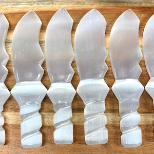 May include: Six selenite crystal knives with curved blades and spiral handles. The knives are a translucent white colour and are arranged on a wooden surface.