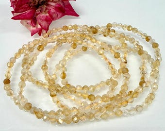 Puede incluir: Un collar de cuentas facetadas, translúcidas, en tonos amarillos y transparentes, dispuestas en forma circular. Las cuentas muestran variaciones de color ámbar y oro pálido, reflejando la luz en sus caras talladas. Una flor rosa oscuro está arriba.