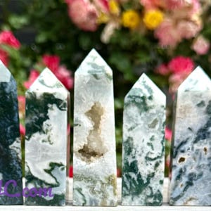 May include: A collection of eight moss agate crystal towers. The crystals vary in color, with green, white, and clear tones. Each tower has a pointed top and a polished surface. The background features a blurred display of pink and yellow flowers.