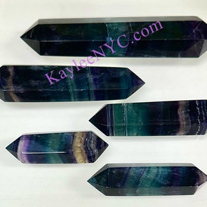 May include: Five double-terminated fluorite crystal points in shades of green, purple, and black. The points are arranged in a staggered pattern on a white surface. KayleeNYC.com is visible in the image.