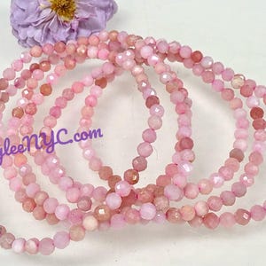 May include: Several strands of pink gemstone beads are arranged in a circular pattern. The beads have a faceted cut, creating a sparkling effect. A purple flower is in the background. The text "KayleeNYC.com" is visible.