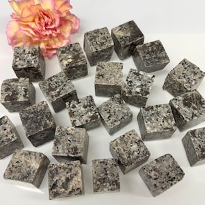 May include: A collection of small, square, gray and white speckled stone blocks. The blocks are scattered on a white surface, with a pink and yellow flower in the background. The blocks appear to be natural stone.