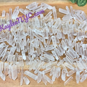 Puede incluir: Una gran colección de puntas de cristal de cuarzo transparente, también conocidas como cristales de roca, esparcidas sobre una superficie de madera. Las puntas son de varios tamaños y formas, algunas son más largas y delgadas que otras.