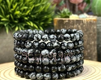 May include: A stack of black and white snowflake obsidian bracelets. Each bracelet is made of round beads with a speckled pattern.