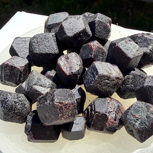 May include: A collection of dark red, faceted garnet stones. The gemstones are piled on a white, shallow dish, with the sunlight highlighting their natural textures and deep color. The garnets vary in size and shape, showcasing their raw, unpolished beauty.