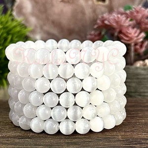Puede incluir: Una pila de cuentas de cristal de selenita blanca, cada cuenta es lisa y redonda. Las cuentas est&aacute;n dispuestas en un patr&oacute;n circular, creando un brazalete o un collar.