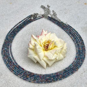 May include: A multi-strand necklace featuring small, faceted beads in shades of blue, purple, and green. The necklace is displayed with a large, creamy white rose with pink edges, creating a striking contrast.