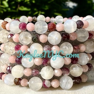 May include: A stack of bracelets made with moonstone, pink, and red beads. The bracelets are layered on top of each other and have a natural, earthy look.